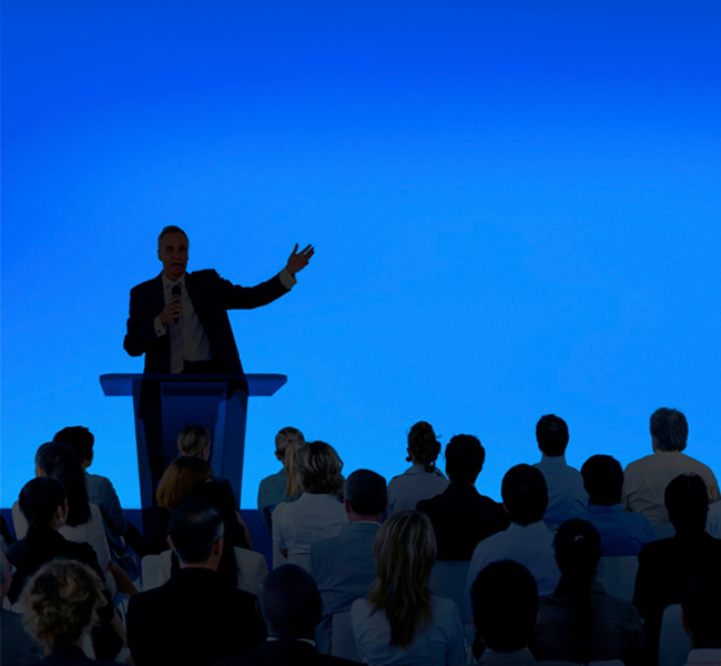 Ejecutivo dando una conferencia ante una audiencia profesional, con fondo azul corporativo