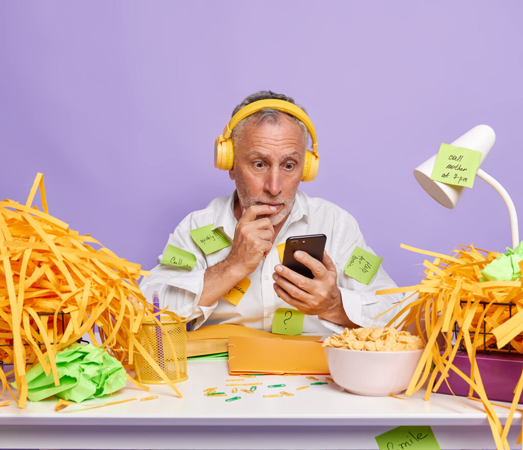 Hombre mayor sorprendido mirando el móvil, rodeado de papeles y notas adhesivas mientras trabaja con auriculares.