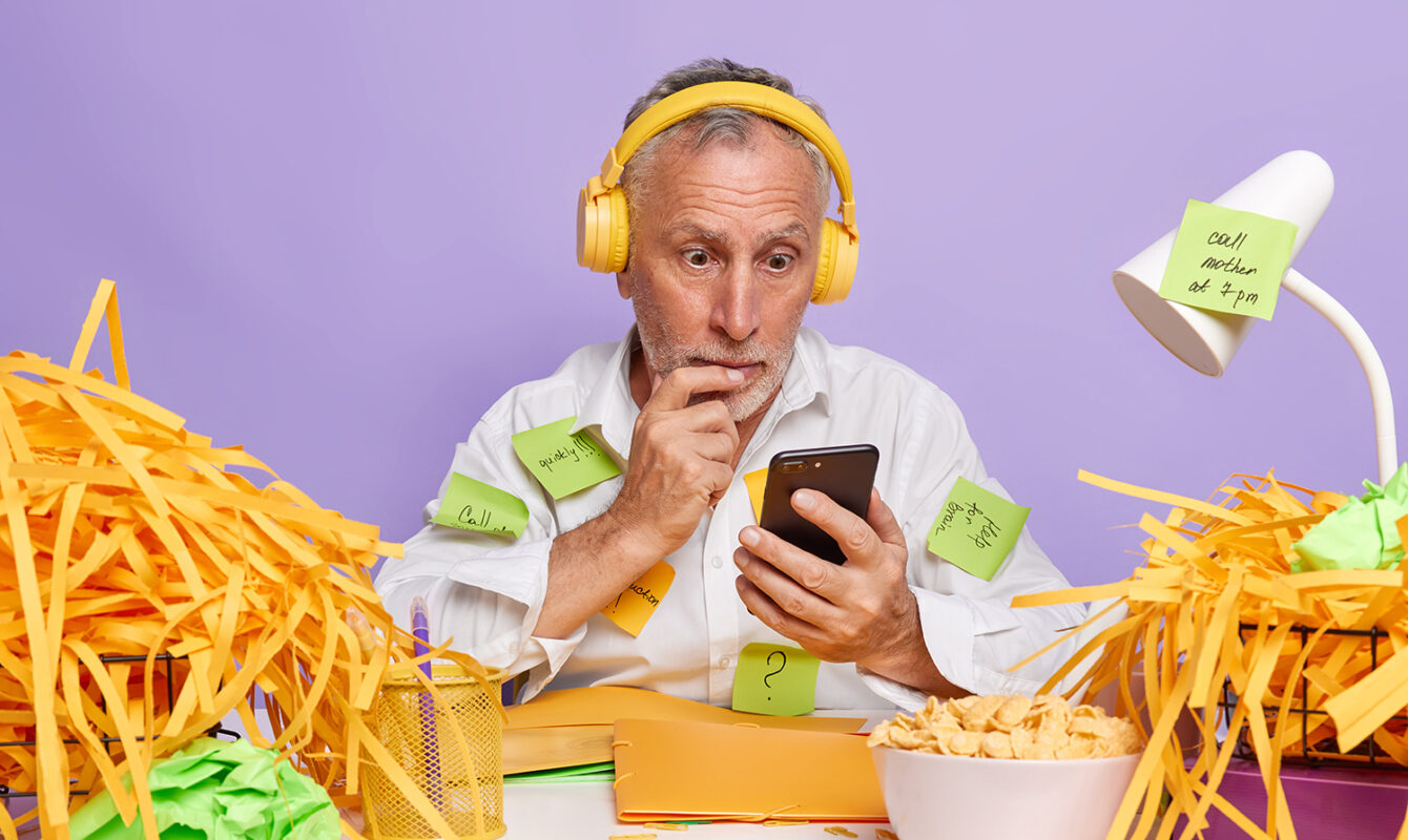 Hombre mayor sorprendido mirando el móvil, rodeado de papeles y notas adhesivas mientras trabaja con auriculares.