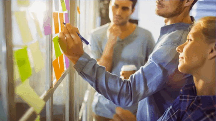 Tres personas colaboran frente a una pared con post-its, escribiendo y organizando ideas en equipo.