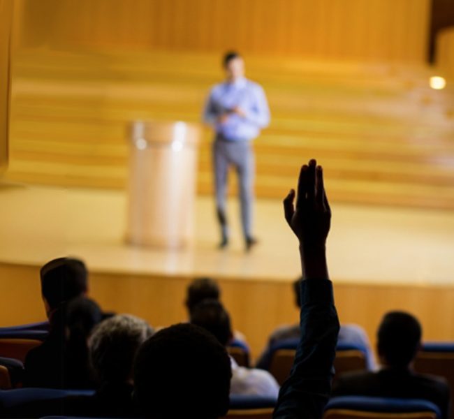 Persona levantando la mano durante una charla en un auditorio con ponente en el escenario