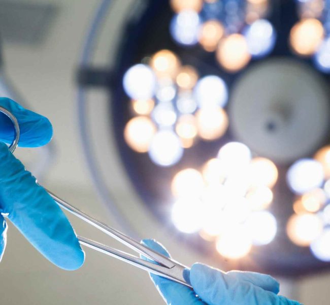 Manos con guantes quirúrgicos sujetando instrumental médico bajo luz de quirófano