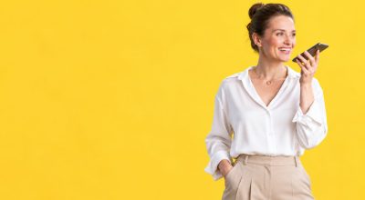 Mujer sonriente con camisa blanca hablando a su smartphone, ilustrando las búsquedas por voz y las nuevas formas de interacción con inteligencia artificial.